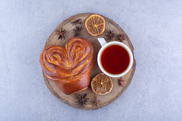 Anis étoilés, tranches de citron séchées, une tasse de thé et un petit pain sucré sur une planche de bois sur une surface en marbre