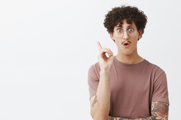 Annonce créative jeune homme intelligent avec moustache cheveux noirs bouclés et bras tatoué levant l'index en geste eureka haletant découvrir une excellente idée ou inventer un plan de génie ajoutant une suggestion