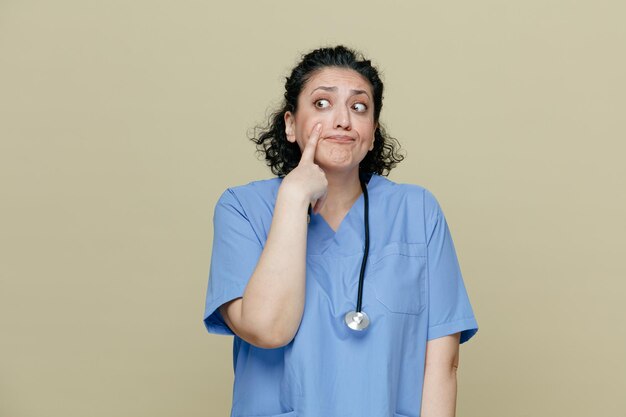 Anxieuse femme médecin d'âge moyen portant un uniforme et un stéthoscope autour du cou regardant le côté pointant le doigt sur le visage isolé sur fond olive