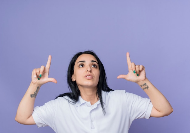 Anxieuse jeune fille caucasienne brune pointe vers le haut avec deux mains isolé sur mur violet