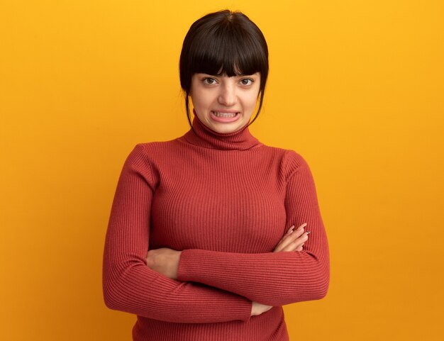 Anxieuse jeune fille caucasienne brune se tient les bras croisés isolés sur un mur orange avec espace pour copie
