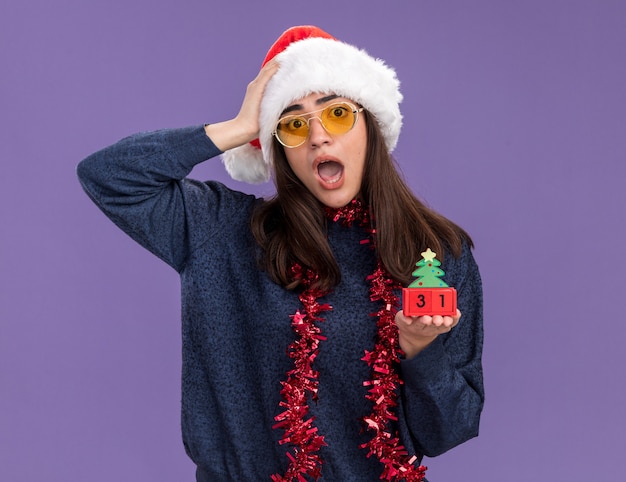 Anxieuse jeune fille caucasienne à lunettes de soleil avec bonnet de noel et guirlande autour du cou met la main sur la tête et tient l'ornement d'arbre de noël isolé sur un mur violet avec espace de copie