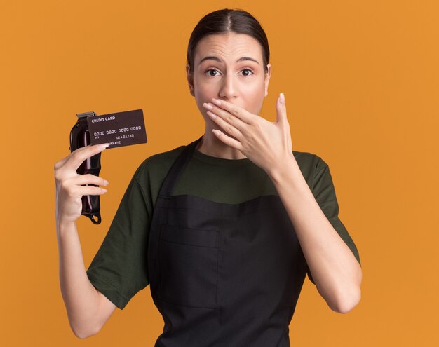 Anxieuse jeune fille de coiffure brune en uniforme tient une tondeuse à cheveux et une carte de crédit mettant la main sur la bouche isolée sur un mur orange avec espace de copie