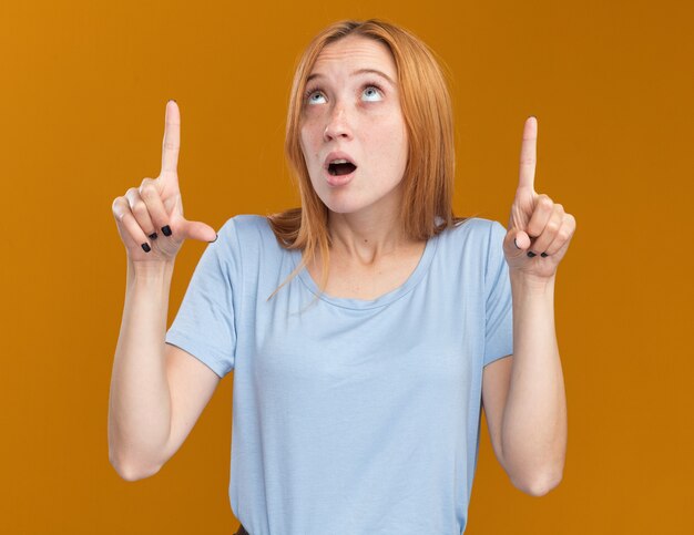 Anxieuse jeune fille rousse au gingembre avec des taches de rousseur regardant et pointant vers le haut avec deux mains isolées sur un mur orange avec espace de copie