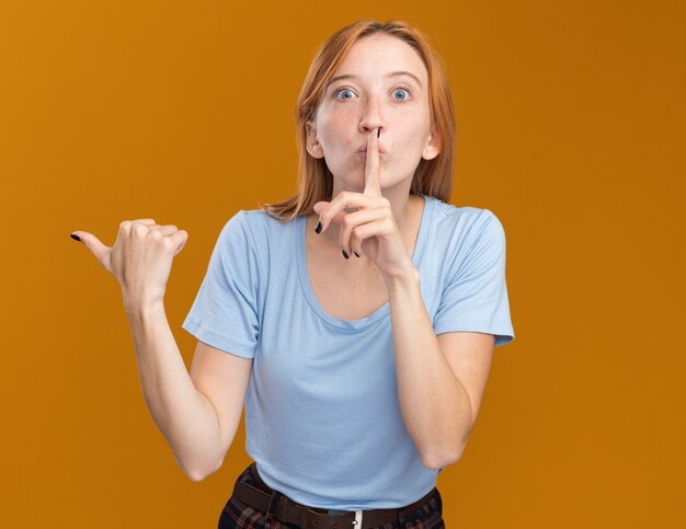 Anxieuse jeune rousse au gingembre avec des taches de rousseur faisant un geste de silence et pointant sur le côté