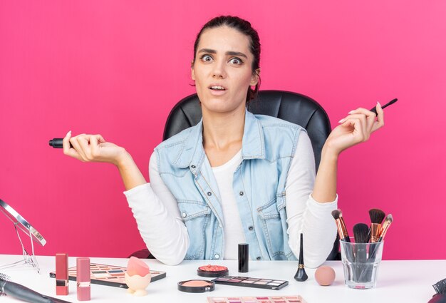 Anxieuse jolie femme caucasienne assise à table avec des outils de maquillage tenant un eye-liner