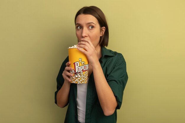 Anxieuse jolie femme de race blanche mange et détient un seau de pop-corn à côté sur vert olive