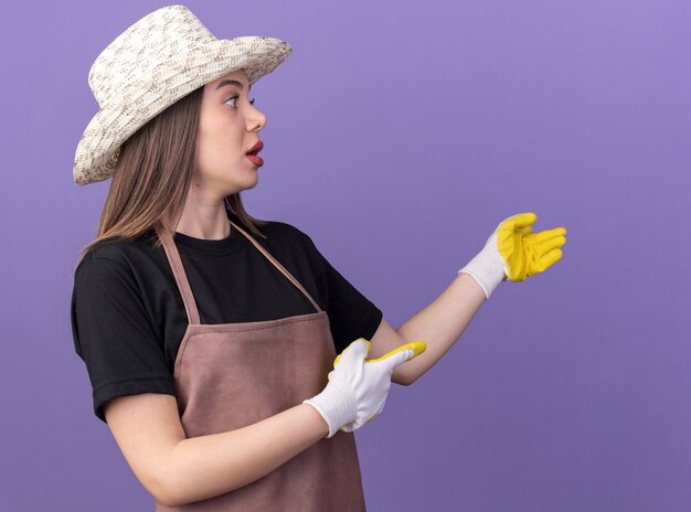 Anxieuse jolie jardinière caucasienne portant un chapeau et des gants de jardinage regardant et pointant sur le côté