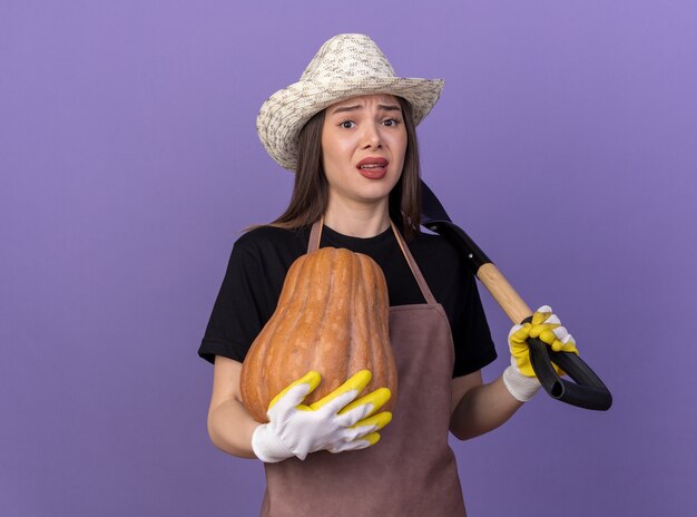 Anxieuse jolie jardinière caucasienne portant un chapeau de jardinage et des gants tenant une citrouille et une pelle sur l'épaule isolée sur un mur violet avec espace pour copie
