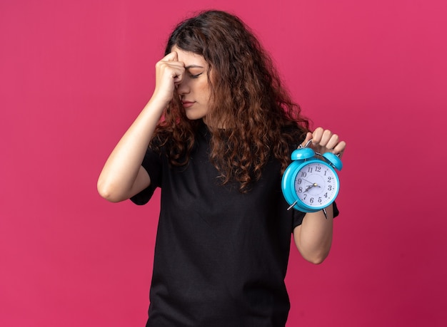 Anxieuse jolie jeune femme gardant la main sur la tête tenant un réveil avec les yeux fermés isolé sur un mur cramoisi