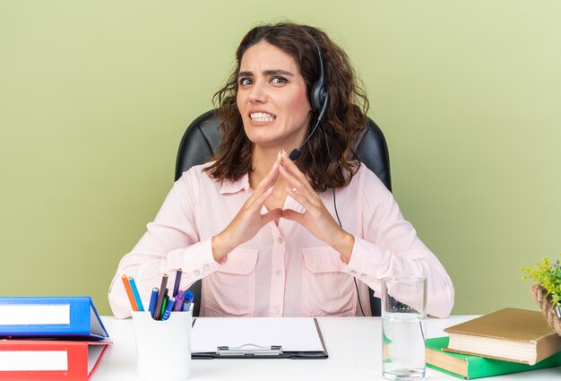 Anxieuse jolie opératrice de centre d'appels caucasienne sur un casque assis au bureau avec des outils de bureau isolés sur un mur vert