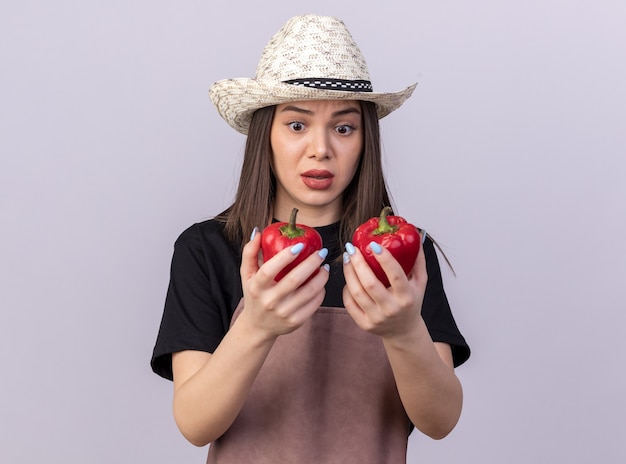 Anxieux assez caucasien femme jardinier portant chapeau de jardinage tenant et regardant les poivrons rouges sur blanc