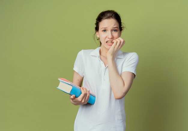 Anxieux jeune étudiante jolie tenant livre et bloc-notes et mordre les doigts isolés sur fond vert olive avec espace de copie