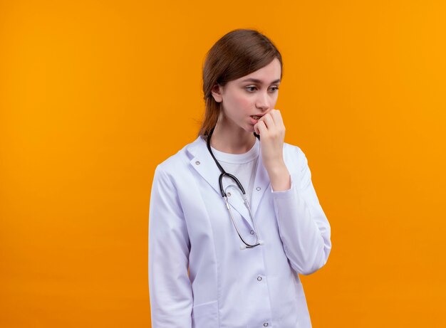 Anxieux jeune femme médecin portant une robe médicale et un stéthoscope et mettant la main sur la lèvre sur un espace orange isolé avec copie espace