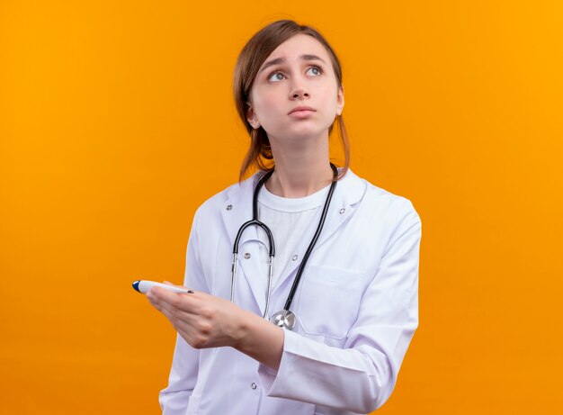 Anxieux jeune femme médecin portant une robe médicale et un stéthoscope tenant un thermomètre sur l'espace orange isolé