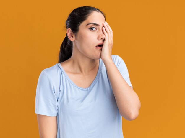 Anxieux jeune fille caucasienne brune met la main sur le visage et regarde la caméra sur orange