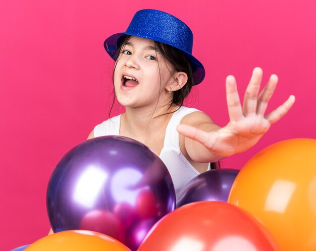 Anxieux jeune fille de race blanche portant chapeau de fête bleu debout avec des ballons d'hélium et étirant la main isolée sur un mur rose avec espace copie