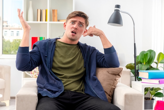 Anxieux jeune homme beau blond est assis sur un fauteuil faisant des gestes signe de la main du téléphone et levant la main à l'intérieur du salon