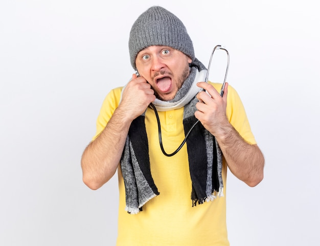 Anxieux jeune homme malade blonde portant un chapeau d'hiver et une écharpe détient un stéthoscope isolé sur un mur blanc