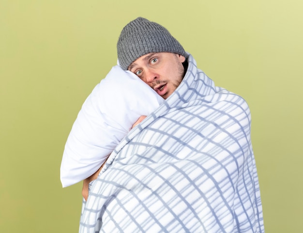 Anxieux jeune homme malade blonde portant un chapeau d'hiver enveloppé dans un oreiller hugs plaid isolé sur mur vert olive