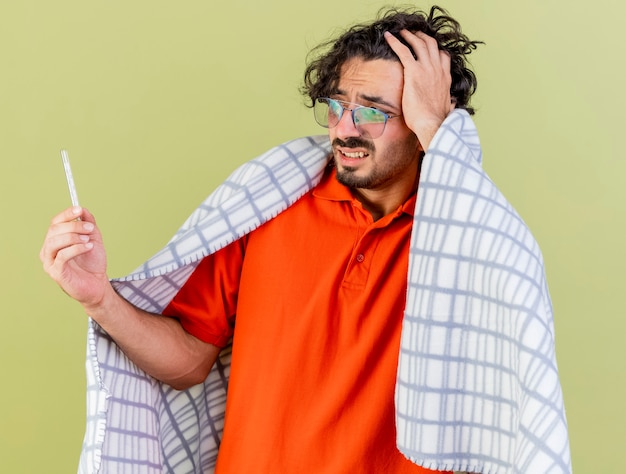 Photo gratuite anxieux jeune homme malade portant des lunettes enveloppé dans un plaid tenant et regardant thermomètre en gardant la main sur la tête isolé sur mur vert olive