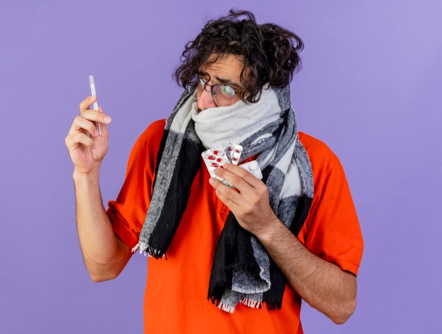 Photo gratuite anxieux jeune homme malade de race blanche portant des lunettes et une écharpe tenant un thermomètre et des pilules médicales regardant thermomètre isolé sur mur violet