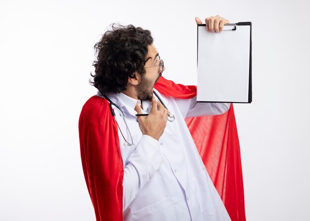 Photo gratuite anxieux jeune homme de super-héros caucasien à lunettes optiques portant l'uniforme du médecin avec manteau rouge et avec un stéthoscope autour du cou regarde le presse-papiers tenant un crayon isolé sur un mur blanc