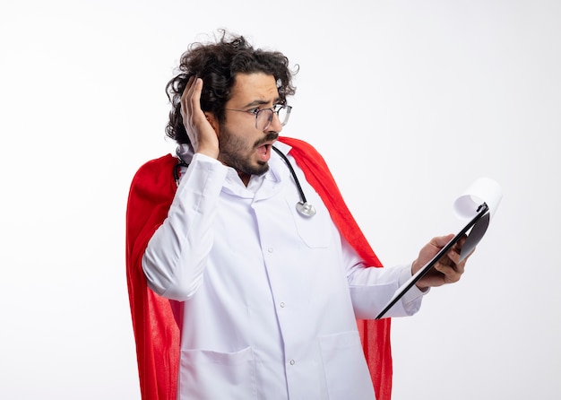 Anxieux jeune homme de super-héros caucasien à lunettes optiques portant l'uniforme de médecin avec manteau rouge et avec stéthoscope autour du cou met la main sur la tête et regarde le presse-papiers avec espace de copie