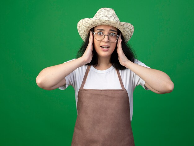 Anxieux jeune jardinière brune à lunettes optiques et uniforme portant chapeau de jardinage met les mains sur le visage isolé sur mur vert