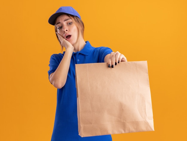 Anxieux jeune jolie livreuse en uniforme met la main sur le visage tenant et regardant le paquet de papier sur orange