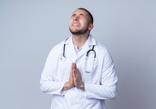 Anxieux jeune médecin de sexe masculin portant une robe médicale et un stéthoscope autour de son cou mettant les mains en geste de prier avec les yeux fermés isolé sur blanc avec espace copie