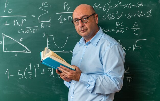 Appareil photo à la recherche d'un enseignant masculin d'âge moyen portant des lunettes debout devant un tableau noir tenant un livre