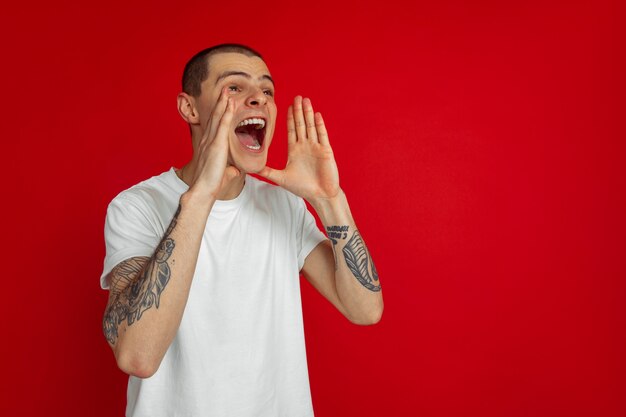 Appel à la vente. Portrait de jeune homme caucasien isolé sur mur rouge. Beau modèle masculin dans un style décontracté. Concept d'émotions humaines, d'expression faciale, de ventes, d'annonces. Espace de copie.