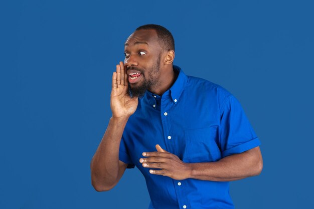 Appeler, crier. Portrait monochrome de jeune homme afro-américain isolé sur le mur bleu du studio.