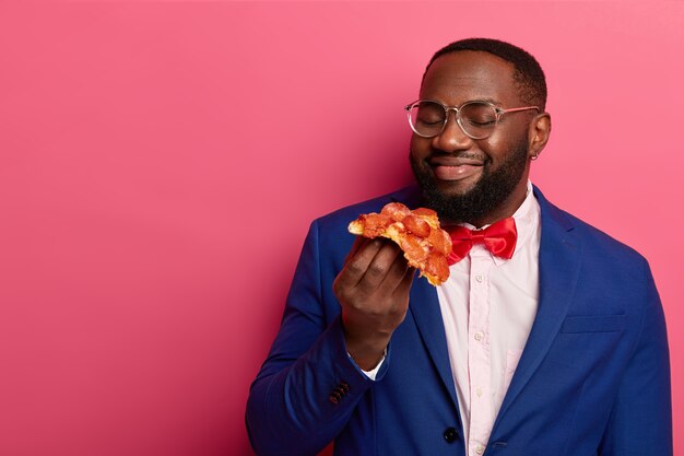 Appétissant de malbouffe. Heureux homme afro-américain à la peau sombre sent la délicieuse pizza, ayant faim après le travail, porte un costume bleu formel, un nœud papillon rouge, une chemise blanche