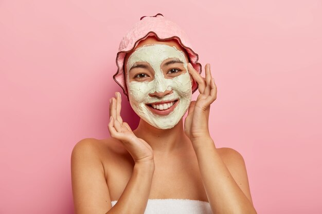 Application de masque facial au spa. Heureuse jeune femme ravie d'apparence asiatique, améliore l'état de la peau avec de la boue d'argile, applique un produit cosmétique sur le visage, prend soin de son teint et de son corps