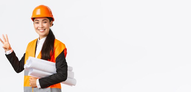 Architecte asiatique souriante dans les domaines de construction de la zone de construction montrant le numéro trois portant le plan directeur comme portant un casque de sécurité donnant un temps fixe pour la tâche effectuée fond blanc