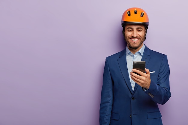 Un architecte joyeux porte un casque orange, un costume formel, a du temps libre pendant la pause de travail, reçoit un message sur smartphone, heureux de recevoir un salaire