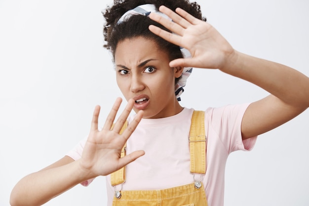 Photo gratuite arrête de l'agiter autour de mon visage. portrait de mécontent et ennuyé femme moderne afro-américaine en salopette jaune et bandeau, couvrant avec des paumes soulevées, fronçant les sourcils