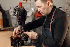 Photo gratuite artisan mature travaillant dans son espace de travail homme portant un tablier et utilisant une machine à coudre fond de texture de pierre sombre grounge