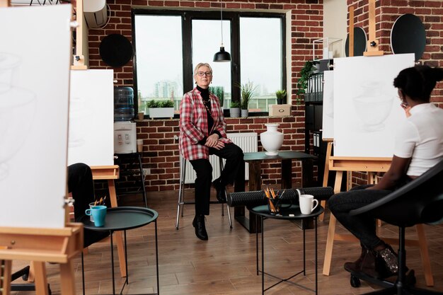 Une artiste âgée enseigne au groupe de personnes à dessiner, assise sur une chaise en classe et souriante à la caméra, atmosphère positive à l'atelier de dessin.