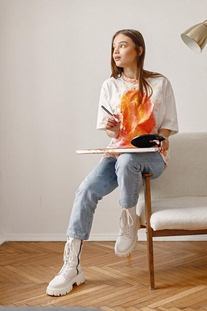 Artiste féminine assise sur une chaise près d'un mur blanc tenant une toile et des pinceaux