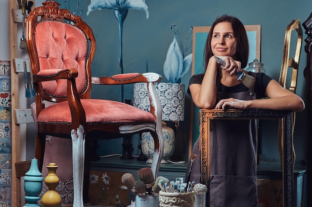 Une artiste féminine pensive en tablier tient un pinceau et s'appuie sur un cadre photo en atelier.