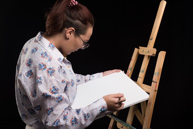 Artiste femme dessinant quelque chose sur toile blanche avec pinceau sur fond noir. Photo de haute qualité