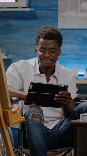 Artiste noir assis en fauteuil roulant à l'aide d'une tablette numérique tout en cherchant l'inspiration pour le dessin d'art. Jeune homme invalide d'origine afro-américaine avec des outils d'art concevant un chef-d'œuvre