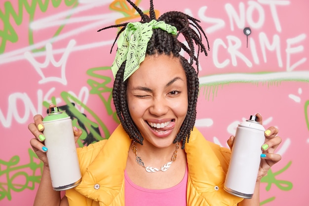 Artiste de rue féminin positif avec une coiffure à la mode cligne de l'œil qui sort de la langue s'amuse tout en peignant un mur de graffitis porte des vêtements clolorful détient des bouteilles aérosol stupides