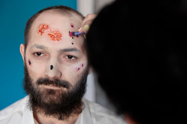 Artiste utilisant le maquillage et le costume pour créer un look zombie, assis en studio. Homme attendant d'avoir l'air dramatique, sanglant et effrayant avec un visage de mort-vivant effrayant et des yeux effrayants. Conception effrayante de cadavre.