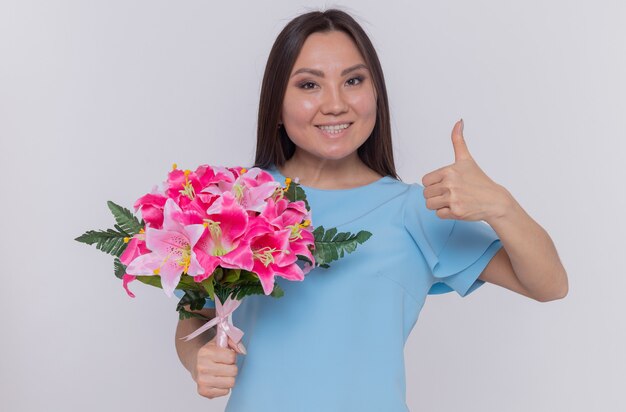 Asian woman holding bouquet de fleurs à la recherche de sourire heureux et gai montrant le pouce vers le haut célébrant la journée internationale de la femme debout sur un mur blanc