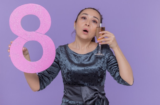 Asian woman holding numéro huit en carton et verre de champagne à la perplexité célébrant la journée internationale de la femme debout sur le mur violet