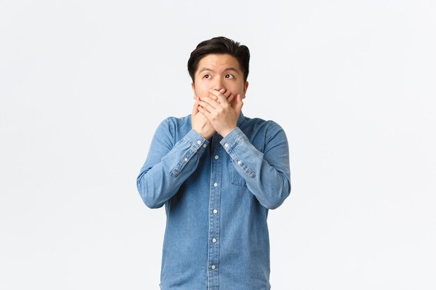 Un asiatique choqué et sans voix est témoin de quelque chose, couvre la bouche avec les mains et regarde dans le coin supérieur gauche, haletant stupéfait par la révélation, entend des potins époustouflants, fond blanc.